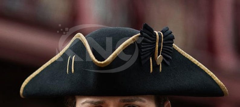 May include: A black tricorn hat with gold trim and accents. The hat features a decorative black bow with gold detailing and a small gold button. The hat's brim is outlined with gold trim.