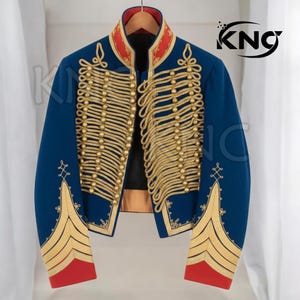 Puede incluir: Chaqueta azul marino de estilo militar con trenzas y botones dorados. La chaqueta presenta cuello y puños rojos, y bordados dorados en las mangas. La chaqueta está colgada en una percha de madera.