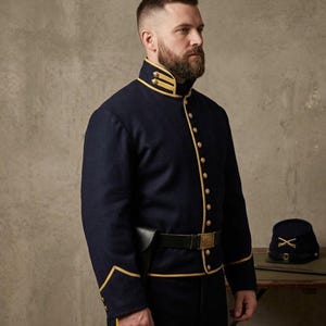 May include: A navy blue military jacket with gold trim and buttons. The jacket has a high collar and is paired with black pants and a belt. A matching hat with gold accents sits on a table. The text "KILT N CRAFT" is visible.