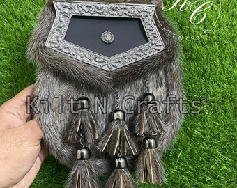 Puede incluir: Un sporran de piel marrón y gris con un adorno de metal plateado y una solapa de cuero negro. El sporran tiene una cadena con cinco borlas.