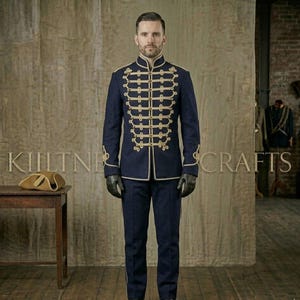 Puede incluir: Un conjunto de estilo militar azul marino compuesto por una chaqueta y un pantalón a juego. La chaqueta presenta adornos y botones decorativos dorados. El conjunto se lleva con guantes negros. Un sombrero color canela está sobre una mesa de madera. El texto "KIILTNER CRAFTS" es visible.