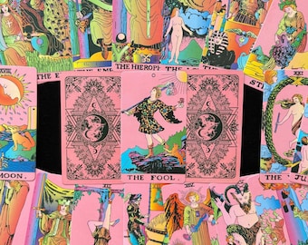 May include: A collection of pink tarot cards with colorful illustrations. The central card features "THE FOOL" with a figure on a mountain. Other cards display various figures and symbols, including "THE MOON" and "THE HIEROPHANT".