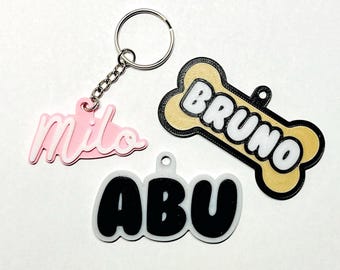 May include: Three personalized dog tags with names "Milo", "Bruno", and "Abu". The tags are made of different materials and colors. The "Milo" tag is pink with white lettering, the "Bruno" tag is a bone shape with a brown wood-like texture and white lettering, and the "Abu" tag is white with black lettering.