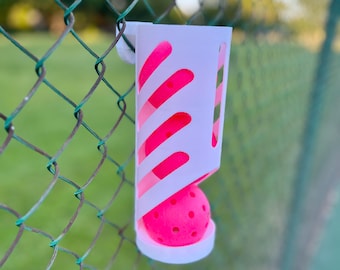 May include: White plastic ball holder with a pink ball inside, hanging on a green chain link fence.