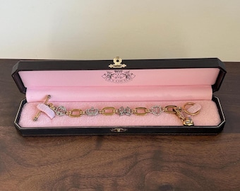 May include: A gold-toned bracelet with various charms, including crowns and hearts, displayed in a black box with a pink interior. The bracelet has a chain-link design and is adorned with rhinestones. The box is open, revealing the jewelry.