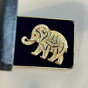 May include: A gold-tone elephant brooch with a red rhinestone eye, covered in clear rhinestones. The brooch is displayed in a black velvet box.