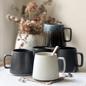 May include: A collection of ceramic mugs in various colors and textures. The mugs include a speckled black, a solid black, a white mug with a wooden spoon, and a mug with a blue and brown glaze. The mugs have large handles and a modern design.