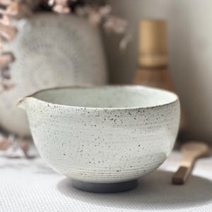 May include: A ceramic bowl with a speckled off-white glaze and a small pouring spout. The exterior features subtle horizontal ridges near the base, which is a solid dark gray color. The interior is smooth and matches the speckled white exterior.
