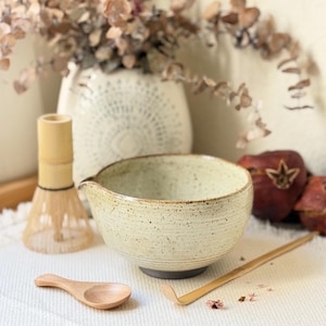 May include: A ceramic matcha bowl with a speckled cream glaze and a small pouring spout, accompanied by a bamboo whisk, a small wooden scoop, and a long, thin bamboo tea ladle. The bowl features a dark base and a textured, rounded shape.