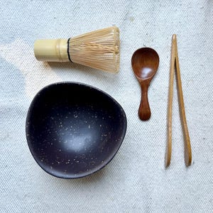 Op de afbeelding: Een matcha theeset bestaande uit een donkerbruine kom met spikkels, een lichtbruine bamboe garde met zwarte draad, een kleine donkerbruine houten lepel en een paar lichtbruine houten tangen op een textuurrijke, gebroken witte stoffen ondergrond.
