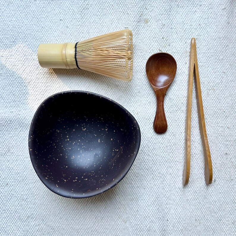 Op de afbeelding: Een matcha theeset bestaande uit een donkerbruine kom met spikkels, een lichtbruine bamboe garde met zwarte draad, een kleine donkerbruine houten lepel en een paar lichtbruine houten tangen op een textuurrijke, gebroken witte stoffen ondergrond.