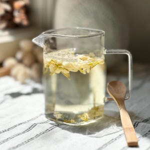 May include: A clear glass pitcher filled with light-colored liquid and floating white jasmine flower buds. The pitcher features a square-shaped handle and a small spout. A light-colored wooden spoon rests against the side of the glass pitcher.