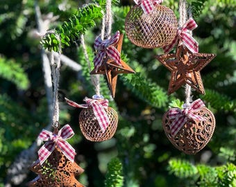 Op de afbeelding: Een verzameling rustieke kerstversieringen hangend aan een tak. De ornamenten, waaronder sterren, bollen en een hart, zijn gemaakt van metaal met een verweerde afwerking. Elk ornament is versierd met een rood-wit geruite strik.