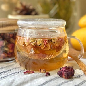 May include: A clear glass tea mug with a matching glass lid containing golden liquid and submerged dried flower petals. A bamboo spoon rests beside the mug, and a glass jar filled with dried rose buds sits in the background near yellow fruit.