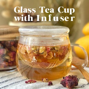 May include: A glass tea cup with an infuser, filled with loose leaf tea. The tea cup has a lid and a handle. The tea is a light amber color and the tea leaves are visible in the water.