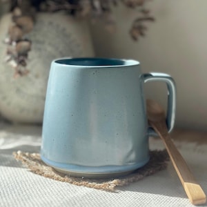 May include: A light blue ceramic mug with a rounded body and a sturdy handle. The mug has a speckled texture and a darker blue interior. A wooden spoon rests beside the mug on a woven coaster. The mug is a handmade item.
