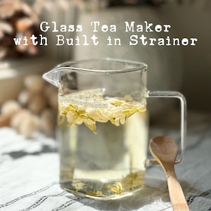 May include: A clear glass tea maker with a built-in strainer, filled with a light-colored liquid and yellow flowers. A small wooden spoon rests on the side. The text "Glass Tea Maker with Built in Strainer" is displayed on the image.