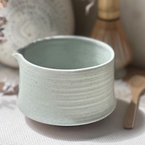 May include: A light green ceramic creamer with a spout, featuring a textured, ribbed design. The creamer is part of a tea set, with a wooden spoon and other tea accessories in the background.