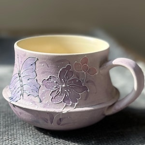 May include: A lavender ceramic mug featuring a raised, white-outlined butterfly and floral design. The mug has a wide, rounded body with a horizontal ridge around the center, a curved handle on the right side, and a smooth, pale cream interior finish.