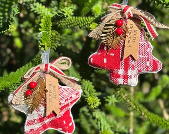 Op de afbeelding: Twee kerstboomversieringen. Eén is in de vorm van een ster, de andere een boom. Beide zijn rood en wit met een ruitpatroon. Elk heeft een strik, bessen en een label met "Merry Christmas!"