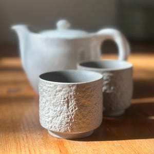 May include: A white ceramic teapot and two textured cups sit on a wooden surface. The cups have a rough, stone-like appearance. The teapot has a curved handle and a lid with a small knob. The set is ideal for serving tea.