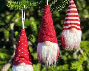 Op de afbeelding: Drie kerstkabouterornamenten hangen aan een tak. Elke kabouter heeft een rode kegelvormige hoed en een witte baard. Eén hoed heeft witte hartjes, een andere is effen rood en de derde heeft rood-witte strepen. De ornamenten hangen aan witte touwtjes.