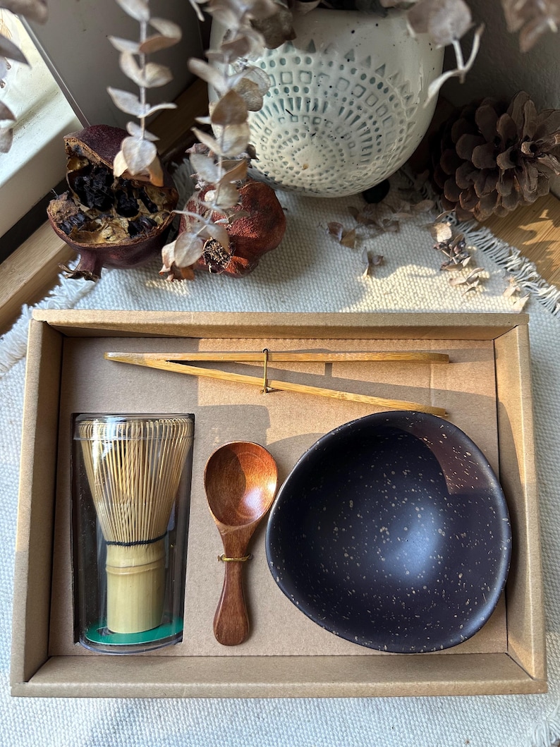 Op de afbeelding: Een matcha-theeset in een kartonnen doos met een bamboe garde in een doorzichtige plastic cilinder, een kleine houten lepel, een dunne bamboe tang en een donkerbruine keramische kom met een gespikkelde afwerking en een onregelmatige rand.