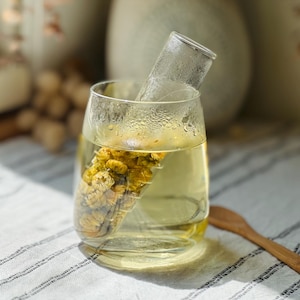 May include: A glass of herbal tea with a clear glass infuser containing dried yellow flowers. The tea is a light yellow color, and the glass has a curved shape. A wooden spoon is in the background.