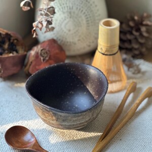 Op de afbeelding: Een donkerbruine keramische kom met een gespikkelde binnenkant en een getextureerde, geribbelde buitenkant aan de basis. De set bevat een kleine houten lepel en een lichte bamboe tang, vergezeld door een bamboe garde achter de kom.