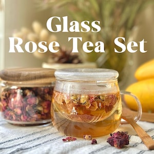 May include: A glass tea set with a clear glass teapot and a glass cup with a handle. The teapot and cup are filled with rose tea. The tea set is on a white and brown striped tablecloth.