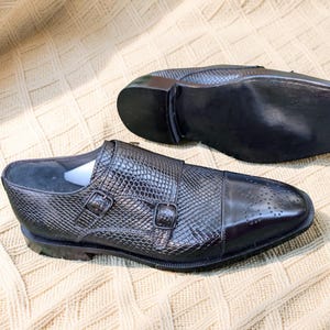 May include: A pair of black double monk strap shoes with a textured snakeskin-patterned upper, a smooth cap toe with subtle brogue detailing, and two matching buckles. The sole of one shoe is visible, featuring a metallic plate at the heel.