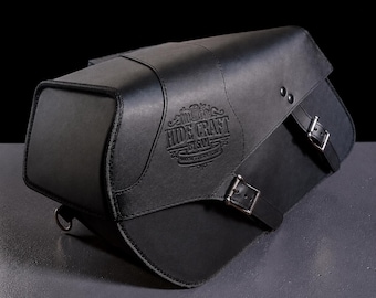 May include: A black leather motorcycle saddlebag featuring a structured rectangular shape with a flap closure secured by two metal buckles. The front panel displays an embossed logo that reads HIDE CRAFT BY SAM with a spool and needle icon.