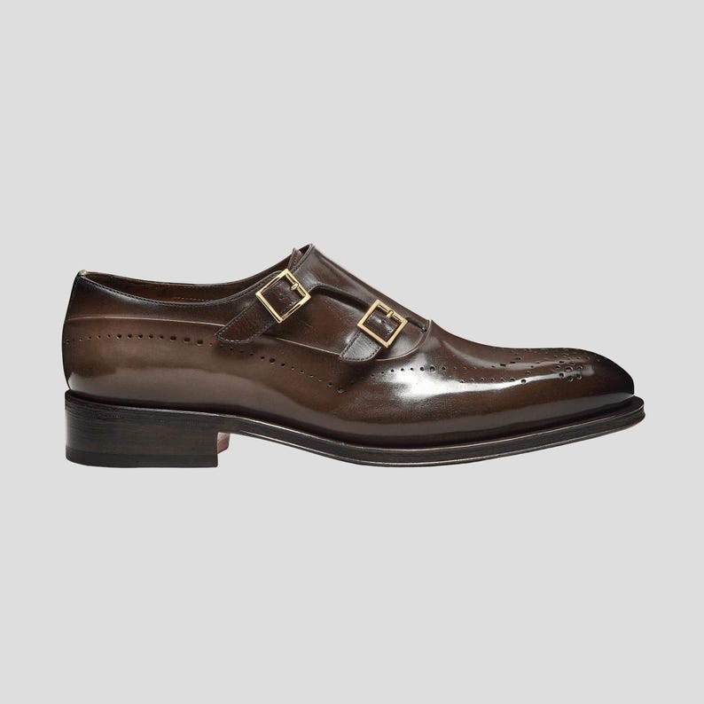Puede incluir: Zapato de vestir de cuero marr&oacute;n con cierre de doble hebilla. El zapato presenta un acabado pulido, perforaciones decorativas y hebillas doradas. La suela es de color marr&oacute;n oscuro con un peque&ntilde;o tac&oacute;n.