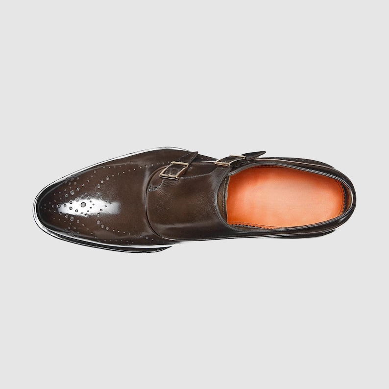 Puede incluir: Vista superior de un zapato de vestir de cuero marr&oacute;n con cierre de doble hebilla. El zapato presenta detalles de tipo brogue en la puntera y una suela de color claro. El interior es de color naranja claro.