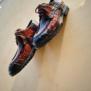 May include: A pair of brown leather dress shoes with a crocodile skin pattern. The shoes have brown laces and a black sole. The leather has a gradient effect, transitioning from a dark brown to a lighter reddish-brown.