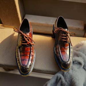 May include: A pair of brown leather dress shoes with a crocodile texture. The shoes have brown laces and a blue trim around the soles. The shoes are displayed on a wooden surface.