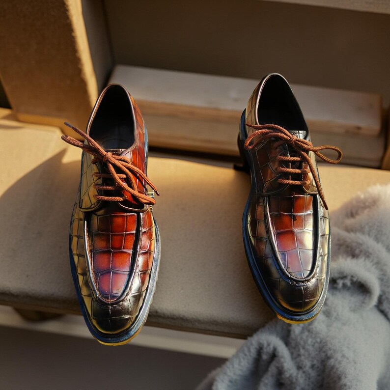 May include: A pair of brown leather dress shoes with a crocodile texture. The shoes have brown laces and a blue trim around the soles. The shoes are displayed on a wooden surface.