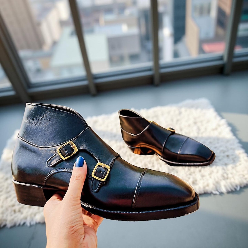 Pu&ograve; includere: Stivaletti in pelle nera con doppie fibbie color oro. Gli stivaletti hanno un design classico con punta arrotondata e tacco basso. Uno stivaletto &egrave; a fuoco, l'altro &egrave; leggermente sfocato sullo sfondo. Gli stivaletti sono su un tappeto bianco.