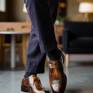 May include: Brown leather dress shoes with a buckle closure and brogue detailing. The shoes have a polished finish and are paired with navy pinstripe trousers and black socks. The shoes are a classic style.