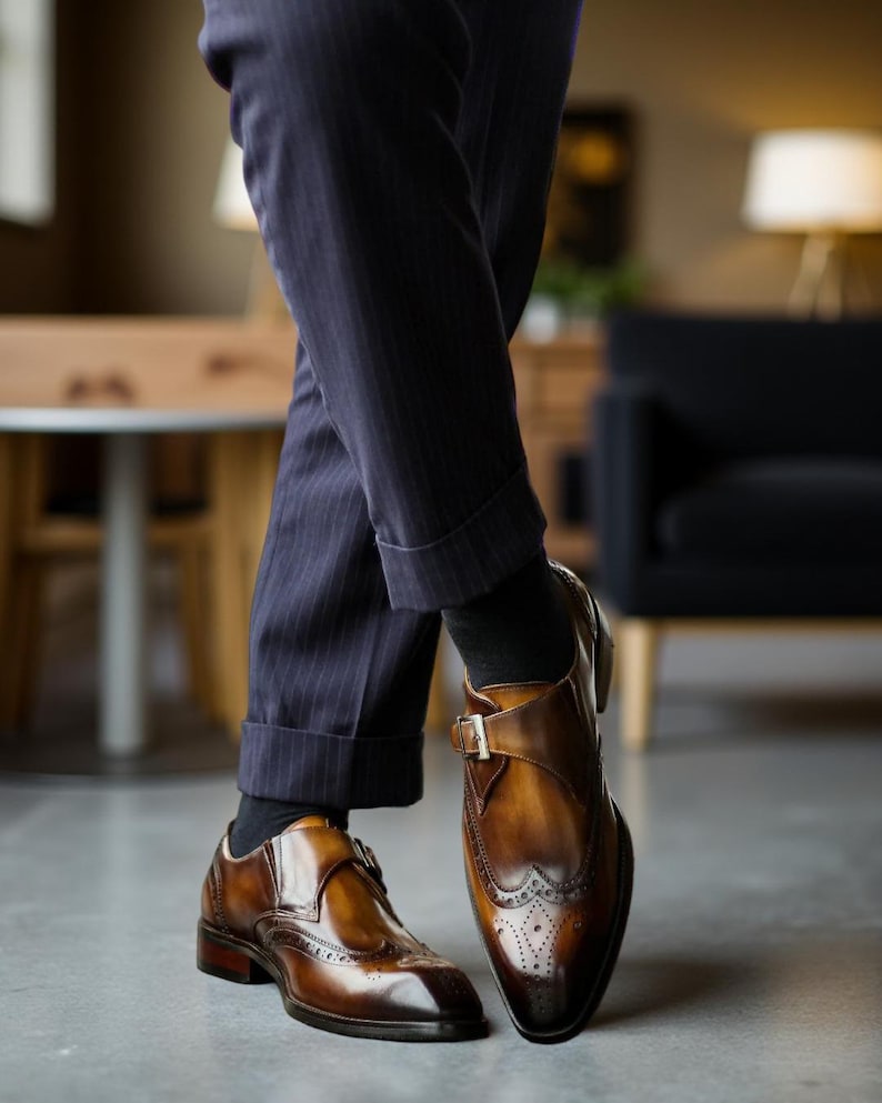 May include: Brown leather dress shoes with a buckle closure and brogue detailing. The shoes have a polished finish and are paired with navy pinstripe trousers and black socks. The shoes are a classic style.