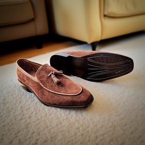 May include: Brown suede loafers with a tassel detail. The shoes have a rounded toe and a contrasting light brown trim. One shoe is upright, while the other is upside down, revealing the sole. The shoes are on a cream-colored rug.