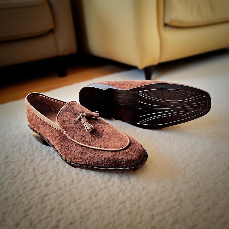 May include: Brown suede loafers with a tassel detail. The shoes have a rounded toe and a contrasting light brown trim. One shoe is upright, while the other is upside down, revealing the sole. The shoes are on a cream-colored rug.