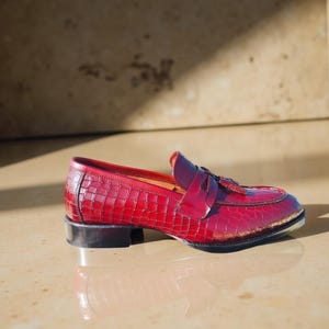 May include: A red leather loafer with a crocodile texture. The shoe has a small heel and a decorative tassel. The shoe is approximately 10 inches long.