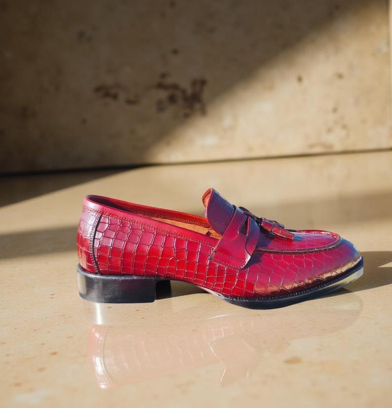 May include: A red leather loafer with a crocodile texture. The shoe has a small heel and a decorative tassel. The shoe is approximately 10 inches long.