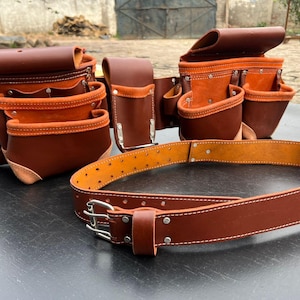 Könnte beinhalten: Ein brauner Leder-Werkzeuggürtel mit mehreren Taschen und einem passenden Gürtel. Die Taschen haben eine orangefarbene Innenseite und sind genietet. Der Gürtel hat eine silberne Schnalle und ist für den professionellen Gebrauch konzipiert.