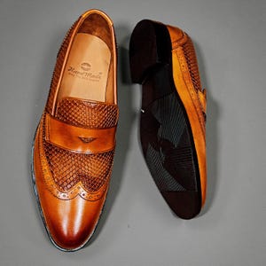 May include: A pair of brown leather loafers with a textured design. The shoes have a classic penny loafer style with a decorative pattern on the vamp and a dark sole. The insole reads "Hand Made Only The Real Leather."