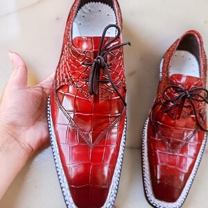 May include: A pair of red leather dress shoes with a textured, crocodile-like pattern. The shoes have a square toe, white stitching around the sole, and dark brown laces. The shoes are displayed on a light-colored surface.