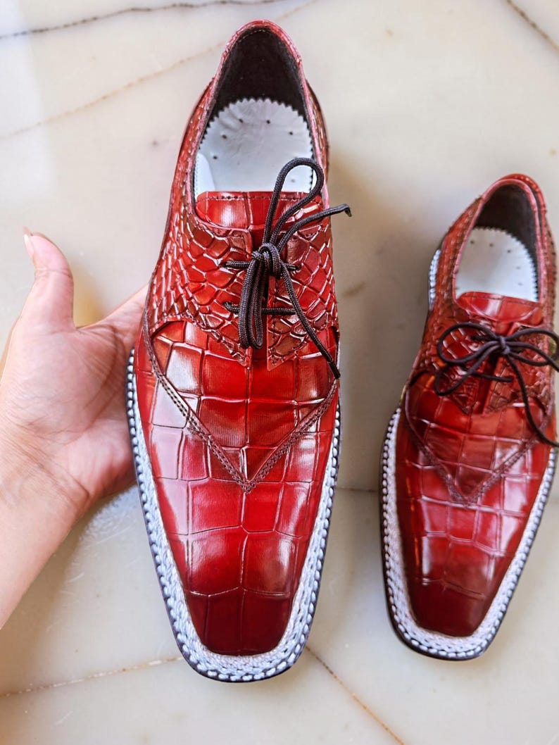 May include: A pair of red leather dress shoes with a textured, crocodile-like pattern. The shoes have a square toe, white stitching around the sole, and dark brown laces. The shoes are displayed on a light-colored surface.