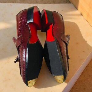 May include: A pair of burgundy leather loafers with a crocodile skin texture. The soles are black with a red accent, and the heels have a gold-colored metal tip. The shoes are displayed with the soles facing up.