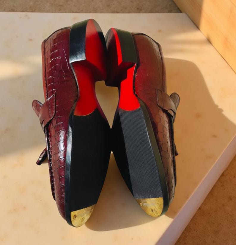 May include: A pair of burgundy leather loafers with a crocodile skin texture. The soles are black with a red accent, and the heels have a gold-colored metal tip. The shoes are displayed with the soles facing up.